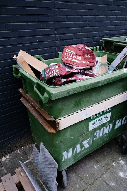 A large green waste bin filled with red and black plastic bags containing construction or gardening materials, placed on a concrete pavement against a dark blue brick wall. The bin has a white label with green text, and a wooden pallet supports it from underneath. The bags are stacked slightly overflowing, with some leaning against the sides of the bin. Nearby, a small metal grate and a few bricks are visible on the ground. The scene suggests private or independent waste removal, typical of a rubbish collection service such as Rubbish Removal Ruislip, operating in a residential or commercial area preparing for rubbish disposal or recycling.