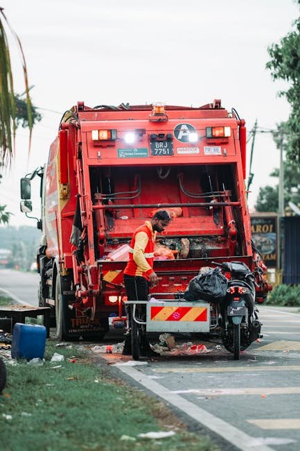 A waste collection worker wearing a yellow high-visibility vest and black trousers is standing at the rear of a large red rubbish collection truck on a roadside. The truck, branded with a white circular logo and identification number BRJ 7751, is positioned on a paved street with a sidewalk and roadside greenery visible. The worker is engaged in emptying or managing a black plastic bag of rubbish, which is placed on a metal platform attached to the truck’s rear. The background includes trees, power lines, and a few commercial signs, with an overcast sky overhead. Nearby, a scooter is parked on the sidewalk, and some scattered waste materials are visible at the foot of the truck. The scene suggests an on-site private waste removal activity consistent with alternative rubbish disposal methods, and the overall environment appears clean and organized, emphasizing the importance of proper waste management services provided by companies like Rubbish Removal Ruislip.